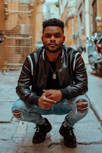 a man crouching down in an alleyway wearing a leather jacket and ripped jeans