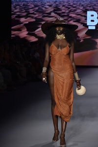 a black woman walks the runway wearing a brown dress and a hat