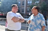 two men in hawaiian shirts talking in front of a fence