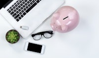 a pink piggy bank, laptop, glasses and a plant on a white table