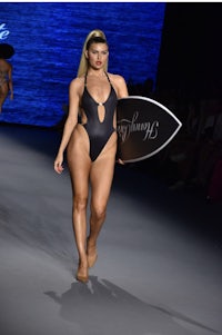 a woman in a black swimsuit walking down the runway with a surfboard